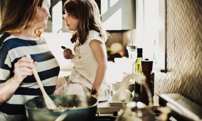 White woman stirring a pot on kitchen stove while smiling at a white child sitting next to her on the counter.